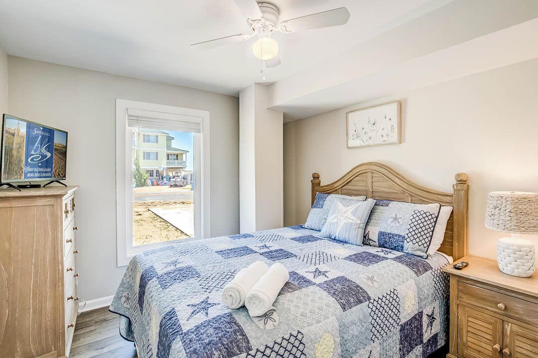 A bedroom with a bed, a ceiling fan, a nightstand, a lamp, and a view of the outside.