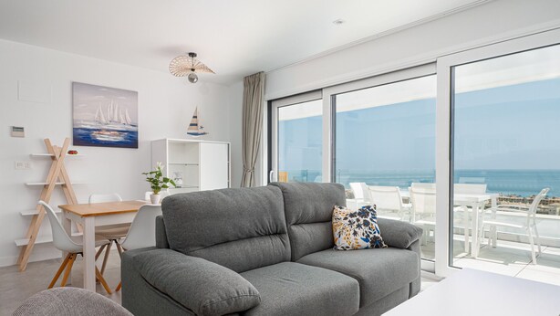 A modern living room with a grey sofa, a white table, and a view of the ocean through large windows.