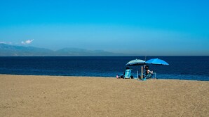 A beach with a clear view of the ocean and distant mountains. There is a blue umbrella providing shade.