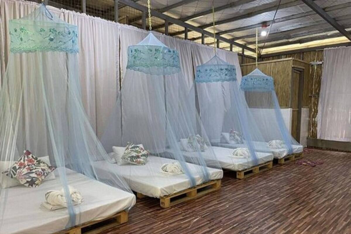 Une rangée de lits protégés par des moustiquaires, des palettes en bois et du linge de lit blanc, dans une pièce au plancher en bois et aux rideaux blancs.