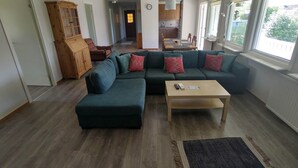 A living room with a sectional sofa, a coffee table, and a rug. There is a wooden cabinet and a dining area in the background.