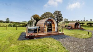 Uma cabana de madeira com hidromassagem e varanda, rodeada por um campo gramado e árvores.
