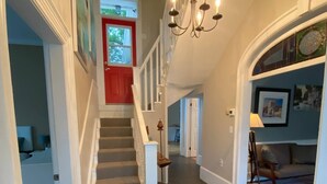 A spacious hallway with a staircase leading to a red door, a chandelier, and a living area with a sofa and a lamp.