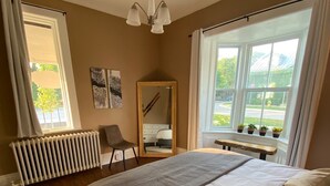 A bedroom with a large bed, a radiator, a chair, a mirror, and a window with plants.