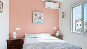 Un dormitorio con cama, mesita de noche, aire acondicionado, ventana con vistas y un cuadro floral en la pared.