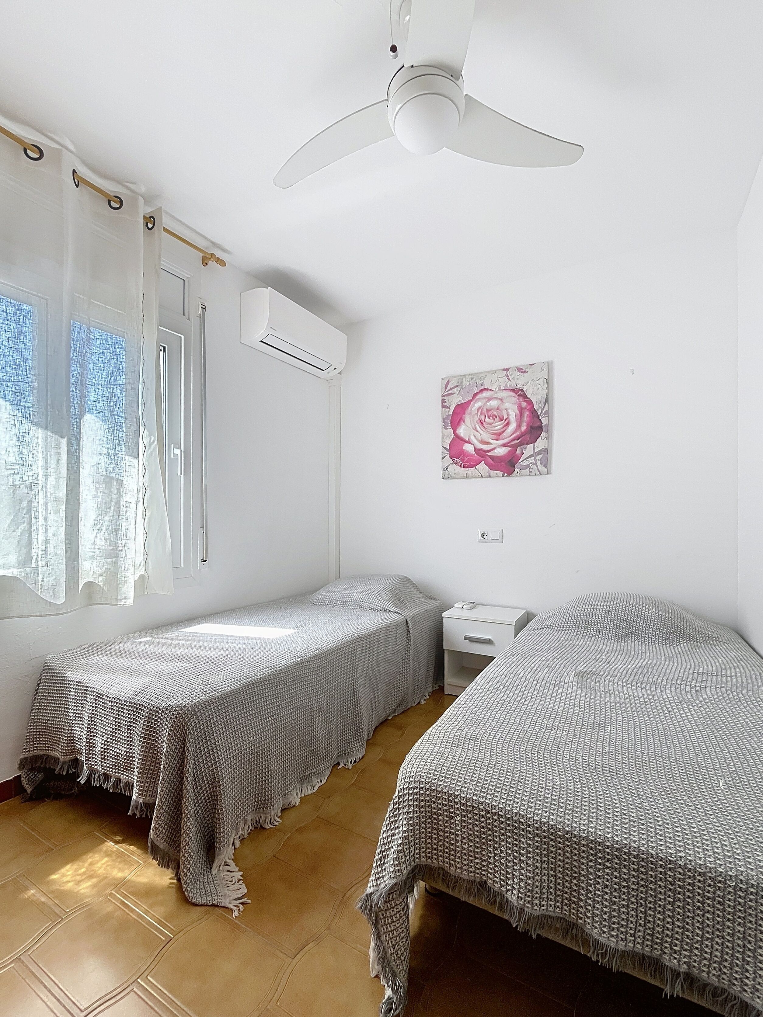 Une petite chambre lumineuse, équipée de deux lits simples, d’un ventilateur de plafond, d’un appareil de climatisation, d’une petite table de chevet et d’une fenêtre avec des rideaux légers.