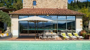 Un espace aménagé au bord de la piscine, avec une table et des chaises, un grand parasol et une vue sur un paysage montagneux, visible à travers de grandes fenêtres.