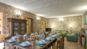 Un espace repas avec une longue table en bois dressée pour un repas, bordé de murs en pierre et agrémenté d’un canapé vert.