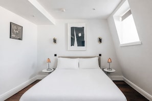 A modern bedroom with a large bed, two bedside tables, a ceiling light, a window, and a wall-mounted abstract painting.