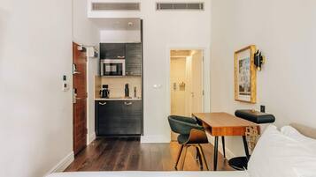 A compact hotel room with a bed, a small desk, a chair, a kitchenette, and a bathroom.