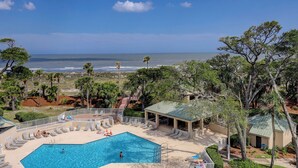 A pool area with a view of the ocean, surrounded by trees and a covered pavilion.