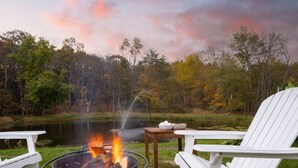 A lakeside campsite with a fire pit, white Adirondack chairs, and a table with bowls.