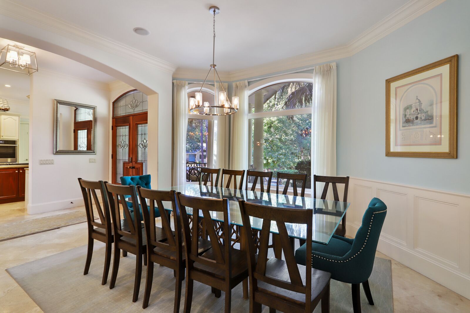 Une salle à manger avec une table en verre entourée de chaises en bois, un lustre et une grande fenêtre avec des rideaux.