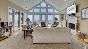 Un salón comedor amplio con techo alto, ventanas grandes que ofrecen vista al océano, chimenea y televisor montado en la pared.