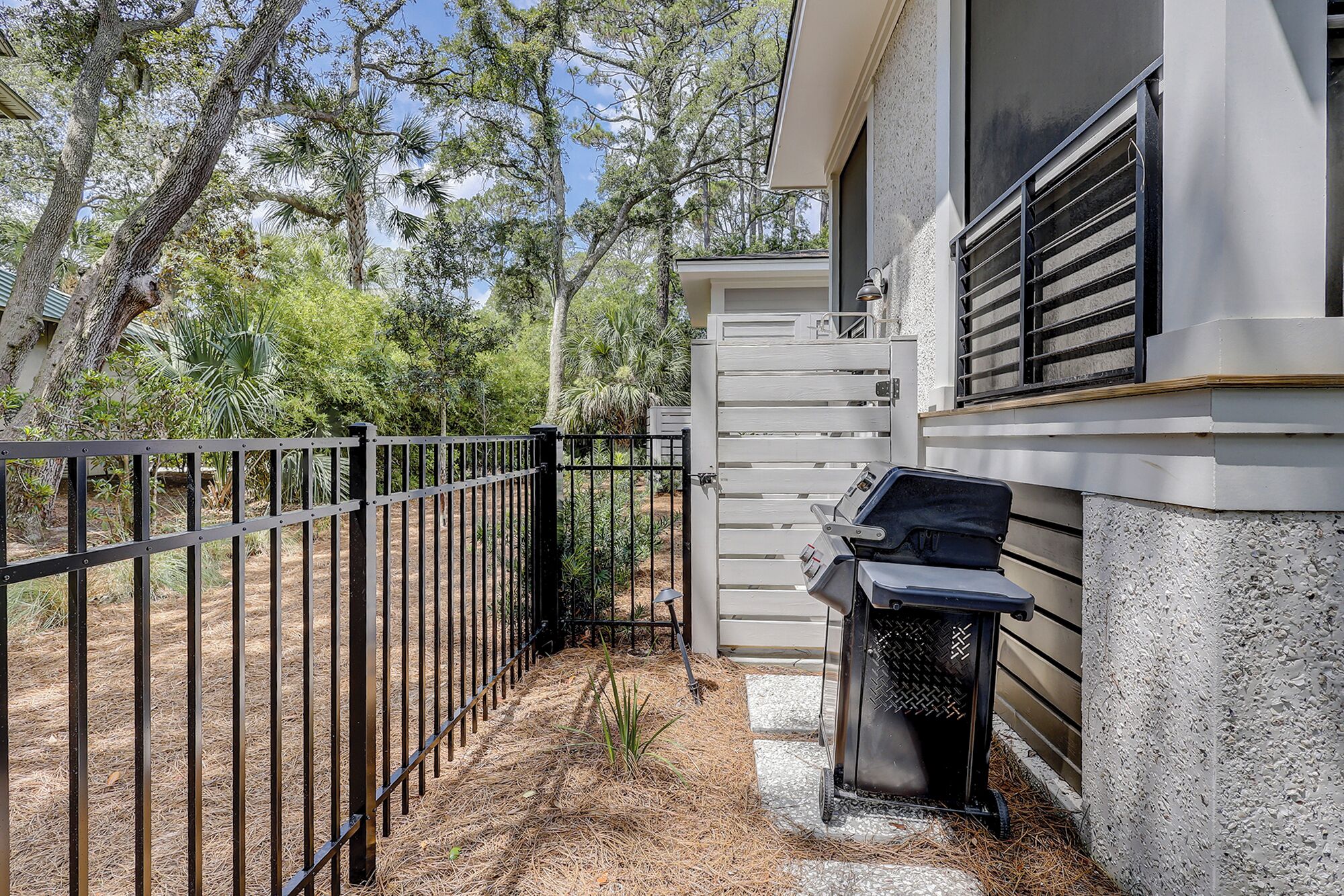 A fenced outdoor area with a grill and a bench, surrounded by trees and a building.