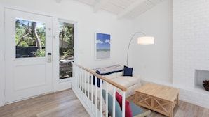 Ein heller Raum mit weißen Wänden und Holzboden, eine weiße Tür mit Blick auf Grünflächen, ein weißes Sofa mit gestreiftem Überwurf, ein Holztisch und eine Stehlampe.