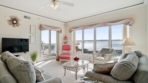 Ein großzügiges Wohnzimmer mit einem großen Fernseher, bequemen Sitzgelegenheiten, einem Balkon mit Aussicht und einem Deckenventilator.