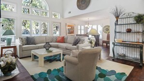 Ein geräumiges Wohnzimmer mit einer großen Eckcouch, einem Couchtisch und einem gemusterten Teppich. Mehrere Fenster sorgen für natürliches Licht, und ein dekorativer Metallregal beherbergt Pflanzen.
