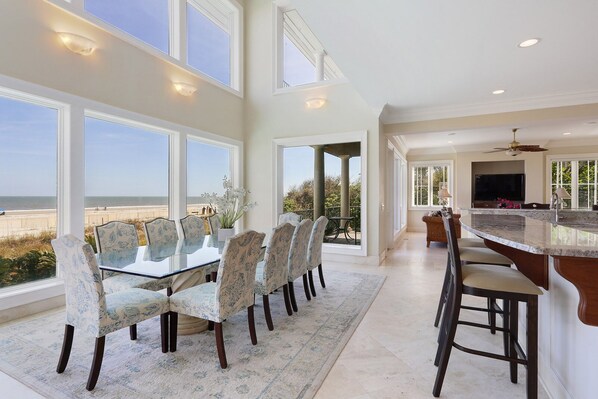 A spacious dining area with a glass table and patterned chairs, overlooking a beach.
