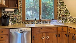 A kitchen with a Bosch dishwasher, wooden cabinets, a large window with a forest view, and a tiled backsplash.