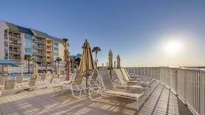 En solbeskinnet dæk med liggestole og parasoller med udsigt over en pool og en hotelbygning.