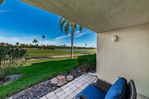 A covered patio with a view of a golf course, a palm tree, and a stone pathway.