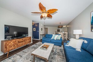 A modern living room with a flat-screen TV, a blue sofa, a wooden cabinet, and a ceiling fan with leaf designs.