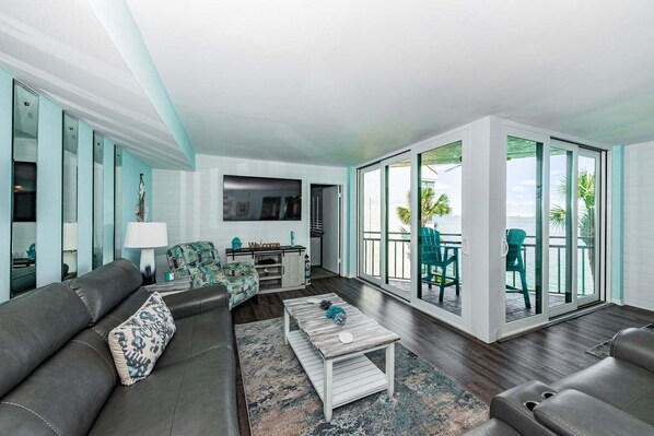 A modern living room with a sofa, coffee table, and chairs, a kitchen area, and a balcony with a view of the ocean.