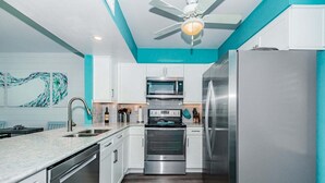 Une cuisine moderne avec des armoires blanches, des appareils en acier inoxydable, un ventilateur de plafond et un mur d’accent de couleur turquoise.