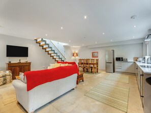 A modern living room with a sofa, a red blanket, a staircase, a dining area with chairs, a kitchen with appliances, and a flat-screen TV mounted on the wall.