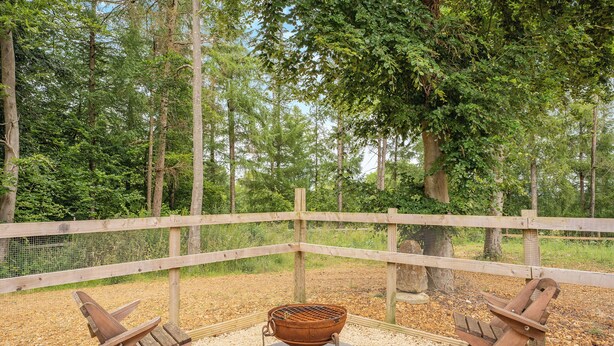 Eine Holzterrasse mit Grilltisch aus Metall und zwei Holzstühlen, umgeben von einem Holzzaun und Bäumen.