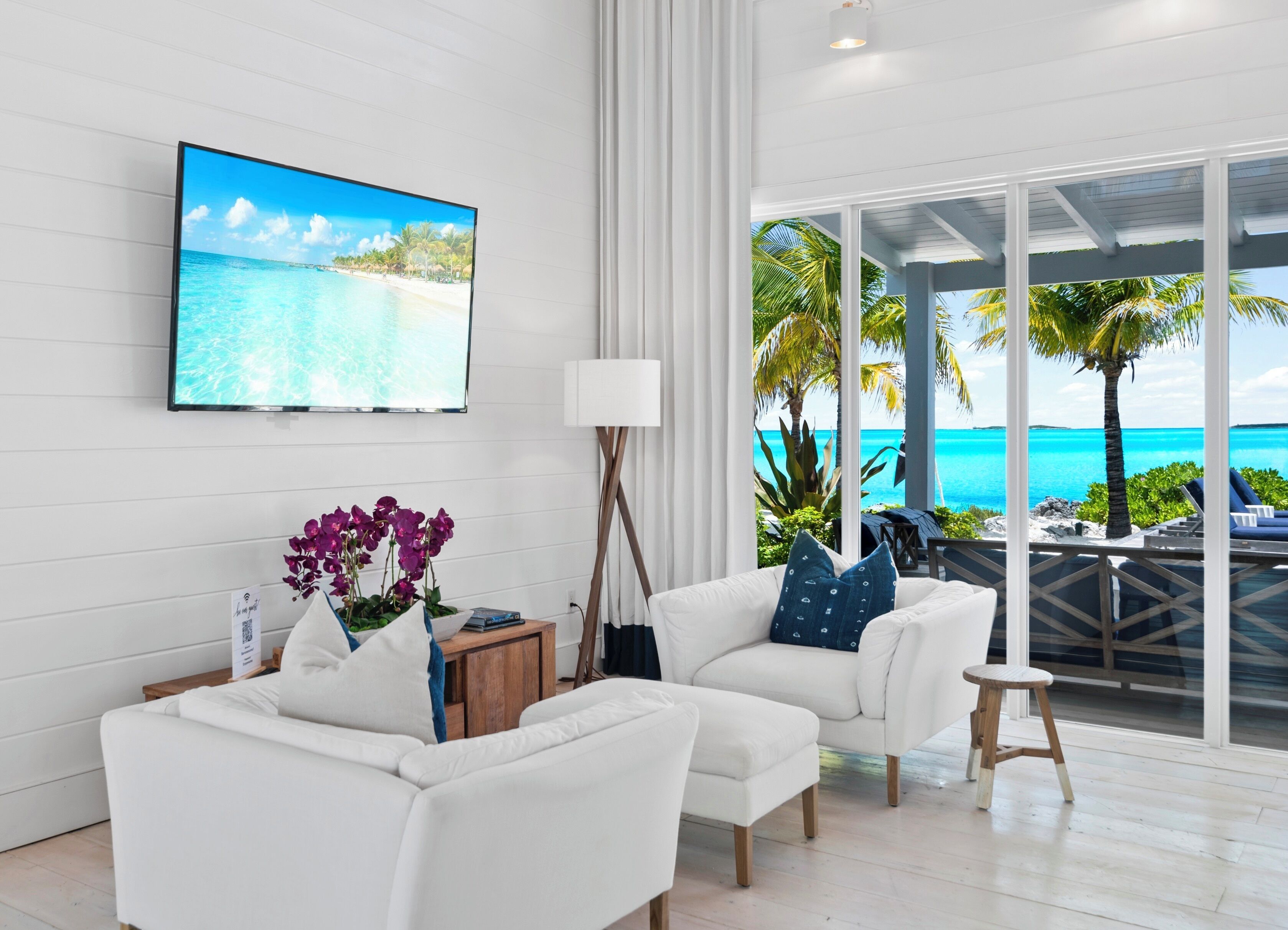 A modern living room with a large TV displaying a beach scene, white sofas, a small wooden side table, and a view of the ocean through large glass doors.