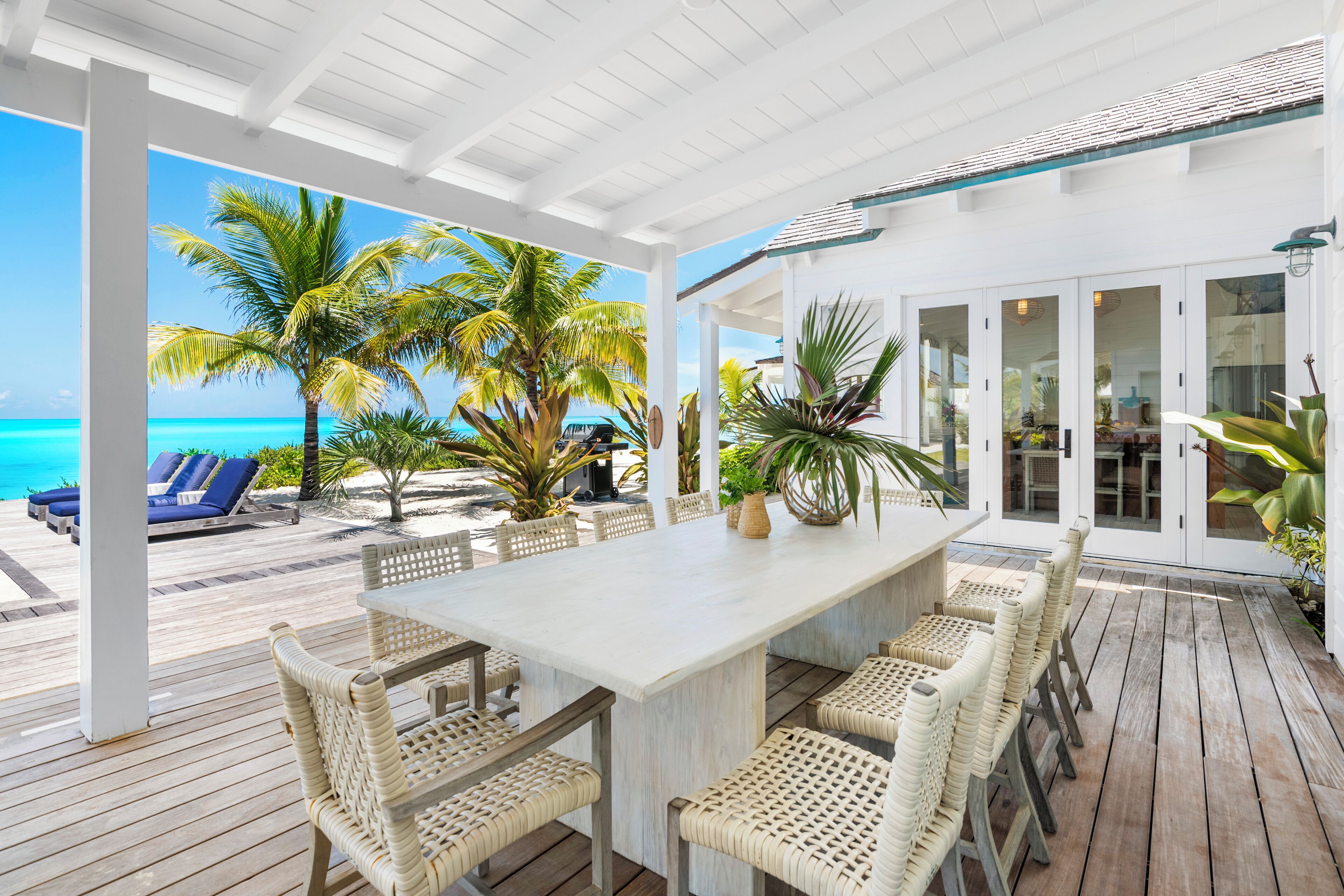Un espace extérieur abrité, aménagé pour les repas, avec une table blanche et des chaises en osier, donnant sur une plage bordée de palmiers et surplombant un ciel bleu azur.