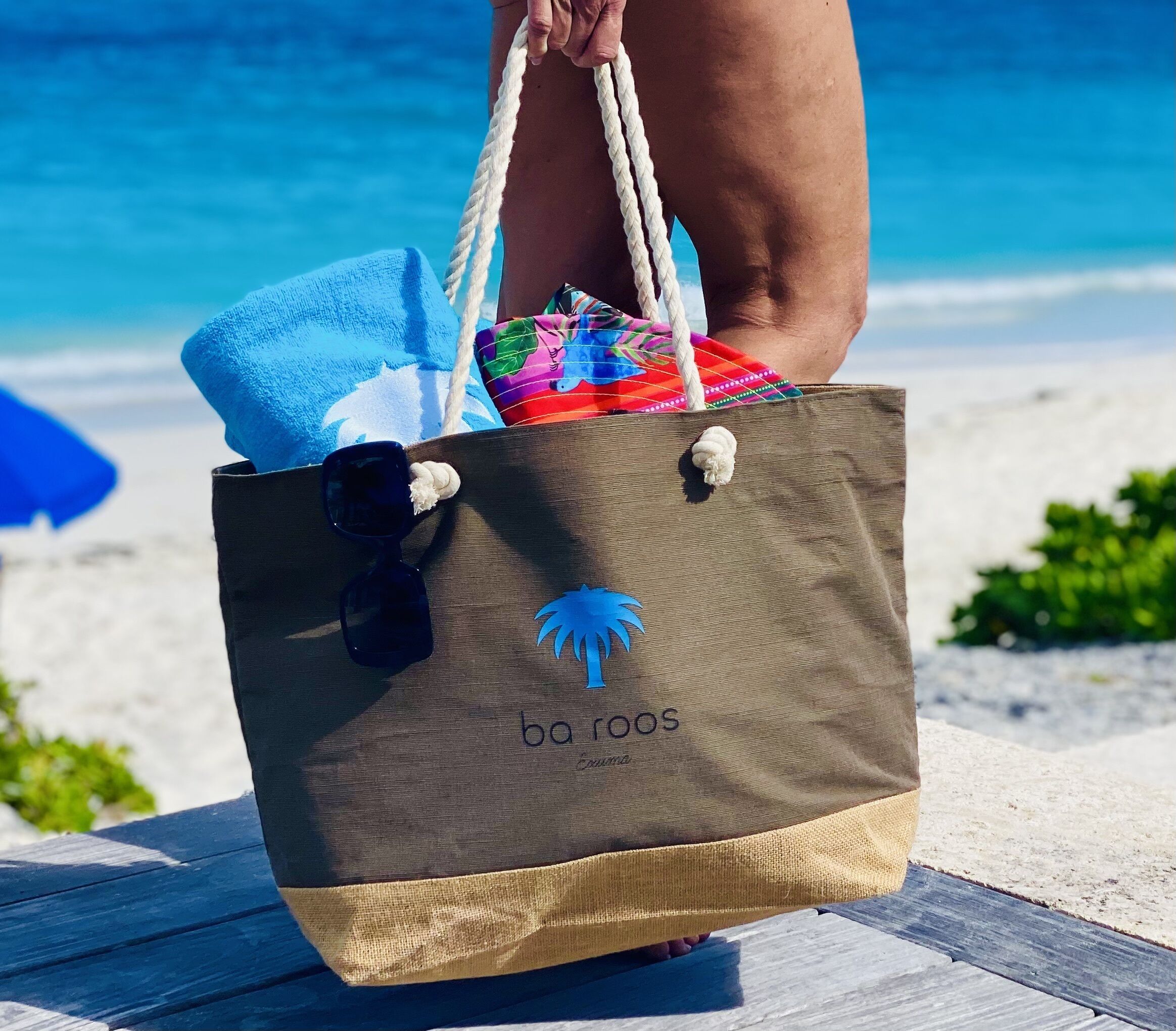 A beachside wooden bench with a blue towel and a colorful bag with a palm tree logo.