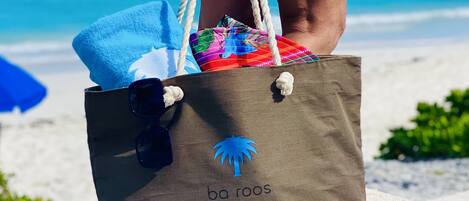 A beachside wooden bench with a blue towel and a colorful bag with a palm tree logo.