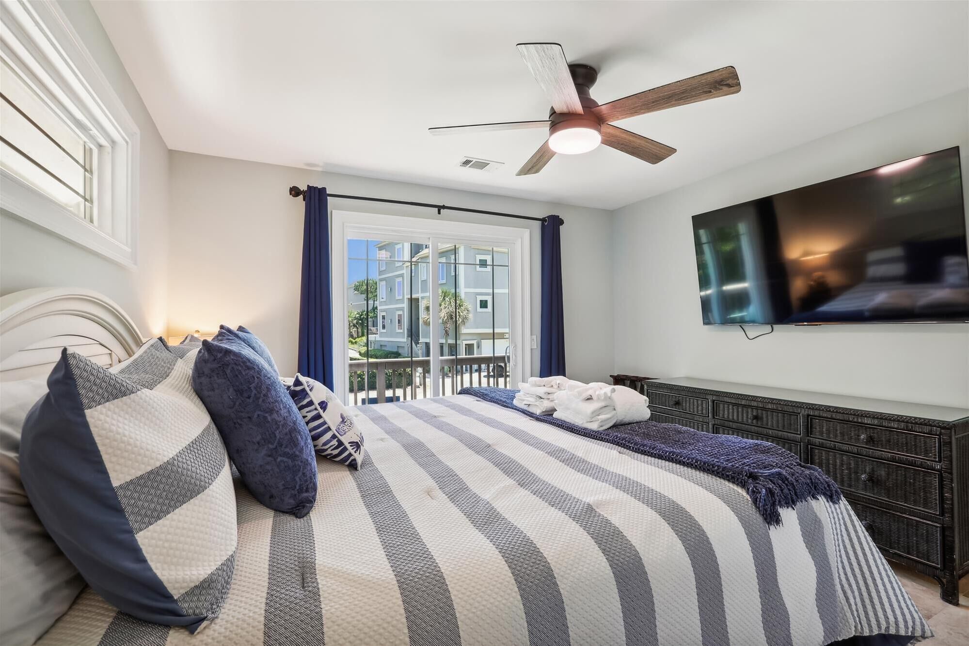 A bedroom with a bed, a ceiling fan, a TV, and a view of buildings outside.