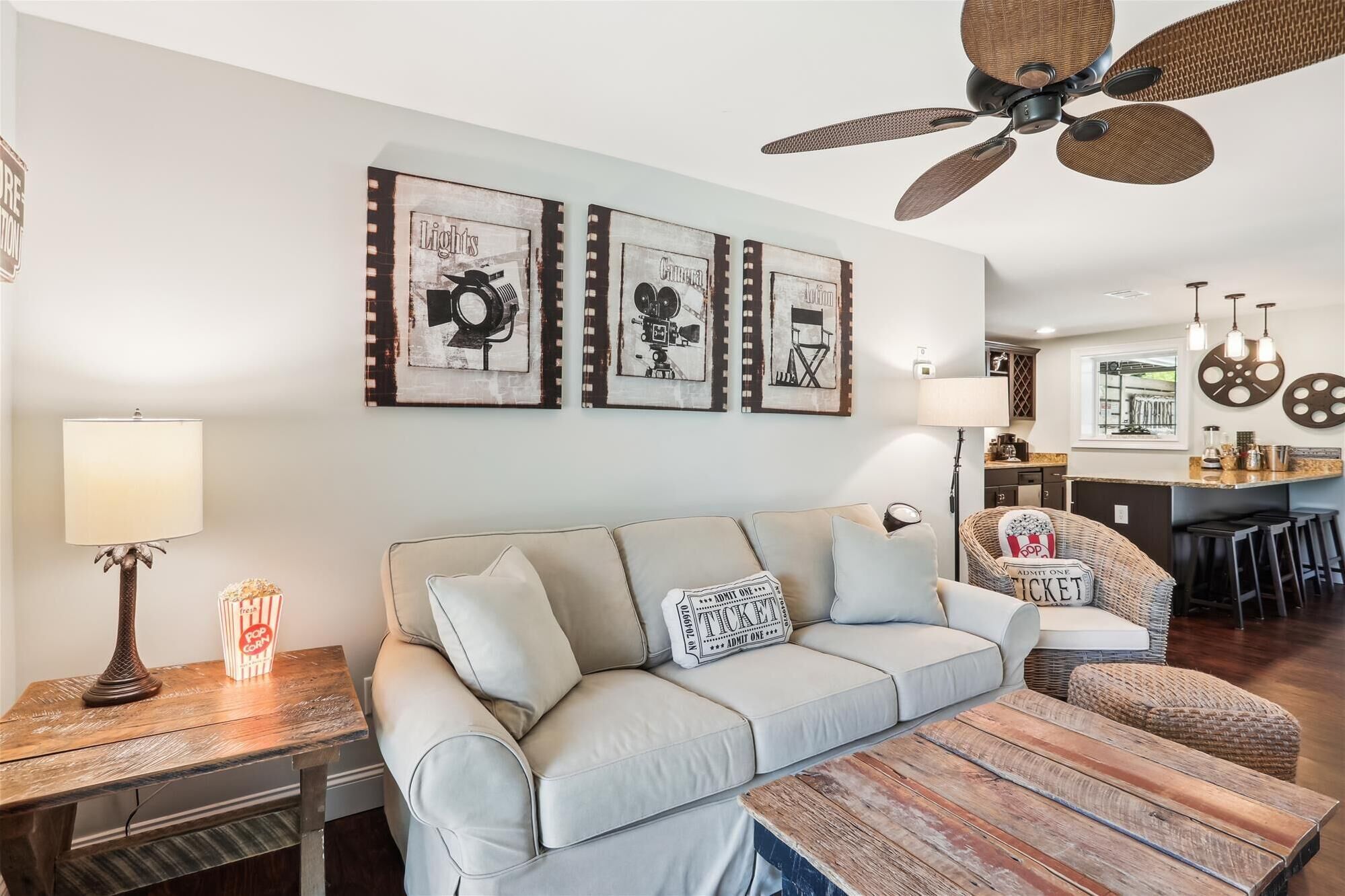 A living room with a sofa, a coffee table, and a dining table. There are framed pictures on the wall, a ceiling fan, and a kitchen area in the background.