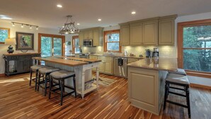 Une cuisine spacieuse dotée d’un plancher en bois, d’un îlot central avec des tabourets et de grandes fenêtres offrant une vue sur l’extérieur.