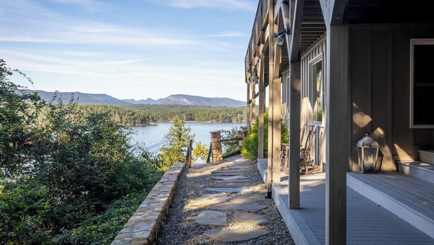 Une terrasse en bois avec un chemin de pierre menant à un bâtiment doté de grandes fenêtres donnant sur un lac et des montagnes.