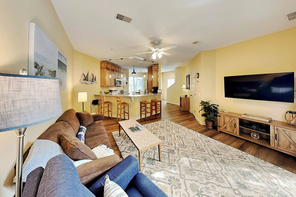 A living room with a sofa, coffee table, and TV. There is a kitchen in the background with a bar and stools.