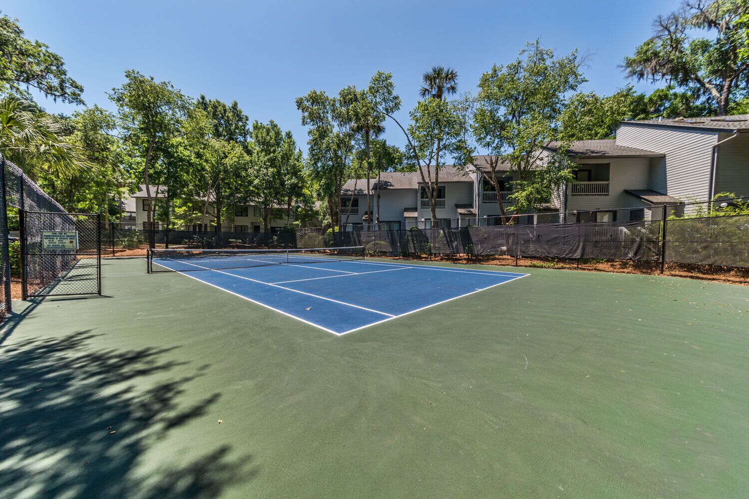 An outdoor tennis court surrounded by trees and residential buildings.