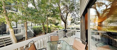A glass-enclosed balcony with a table and chairs, surrounded by trees and a white fence.