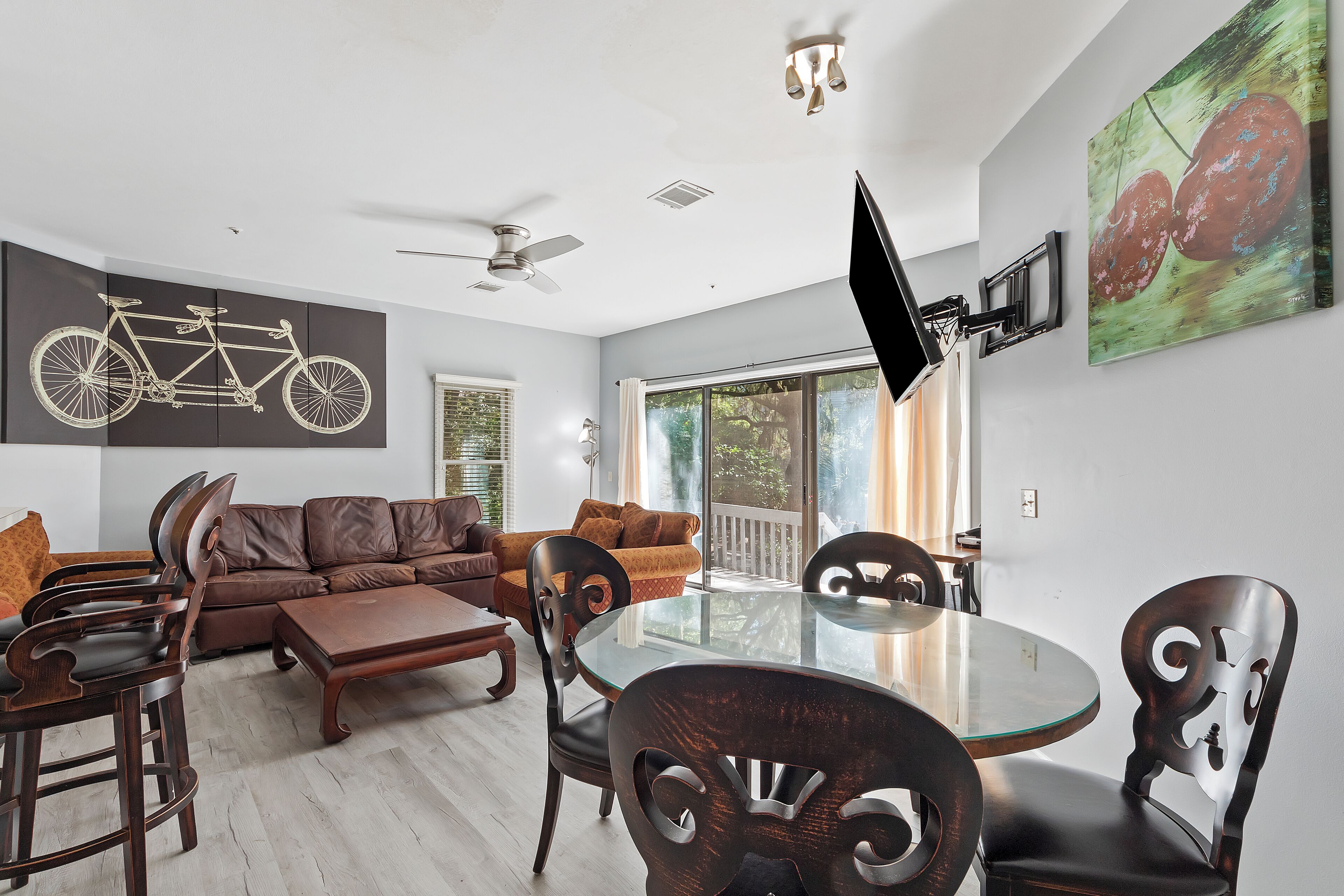 A modern living room with a glass-top dining table, dark wooden chairs, a brown leather sofa, and a ceiling fan.