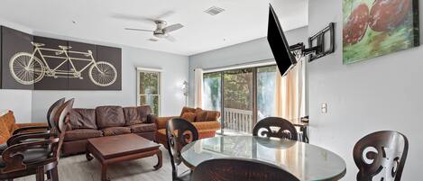 A modern living room with a glass-top dining table, dark wooden chairs, a brown leather sofa, and a ceiling fan.