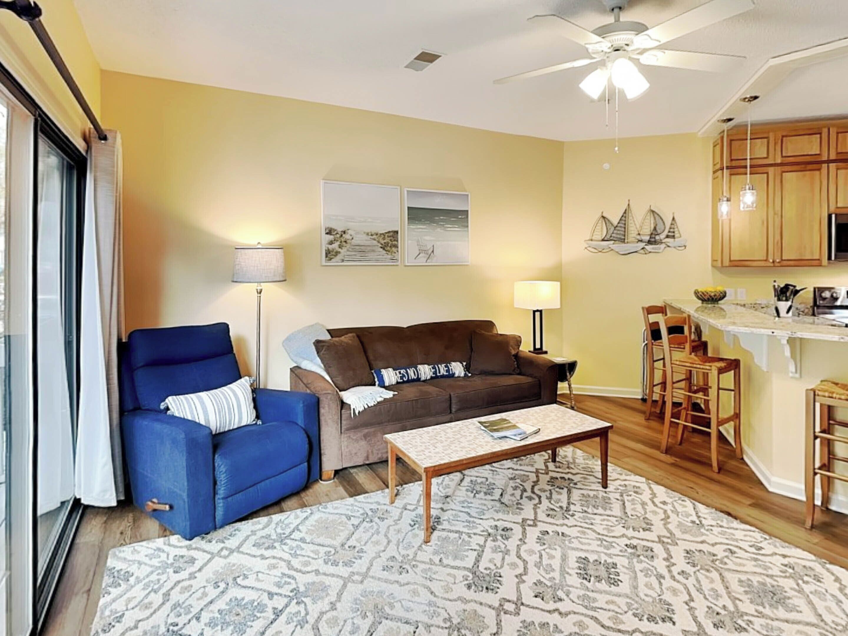 A living room with a blue armchair, a brown sofa, and a coffee table. There is a kitchen area with wooden cabinets and a bar stool.