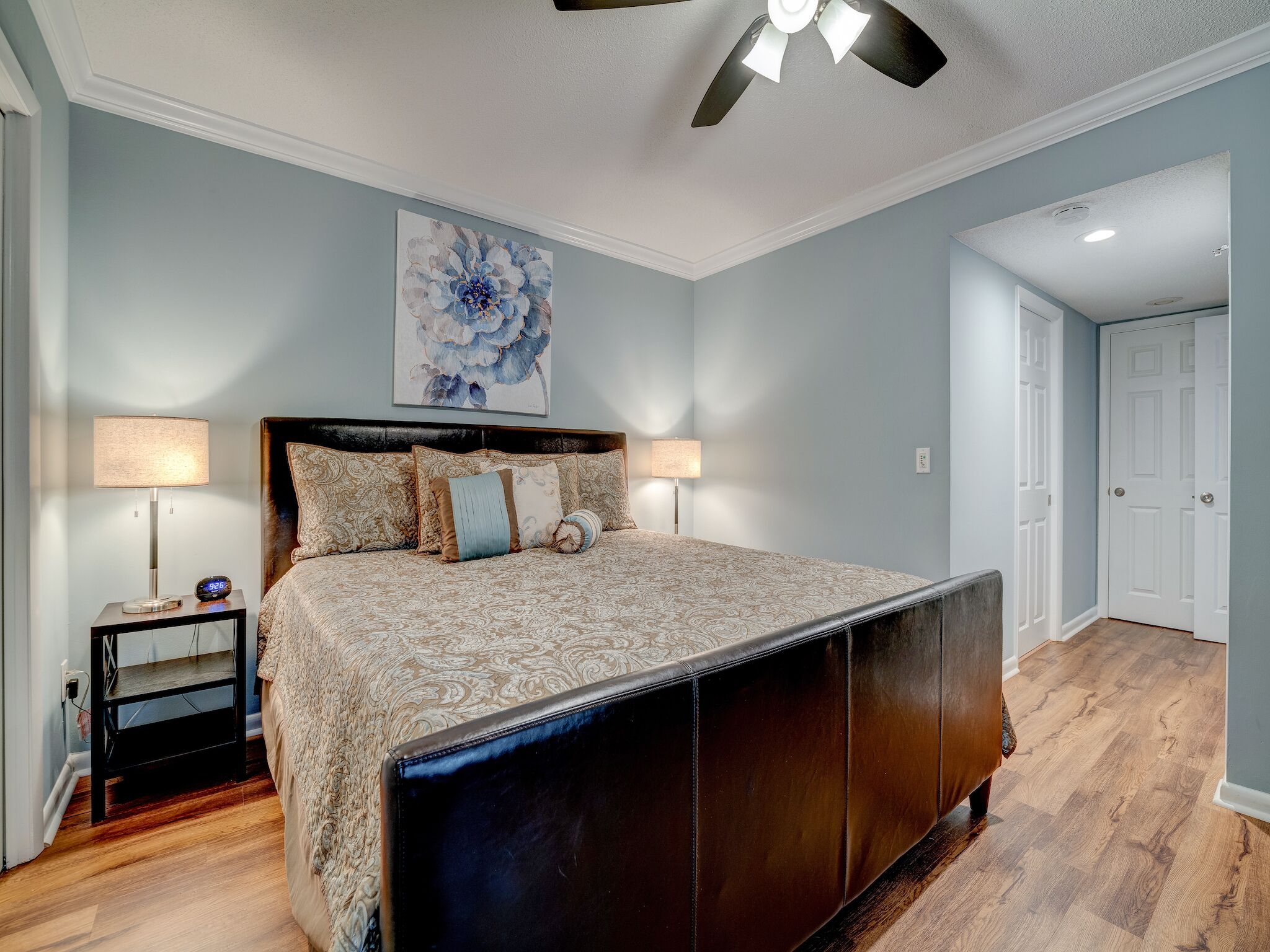 A bedroom with a large bed, two bedside tables, a ceiling fan, and a door leading to another room.
