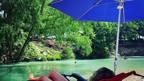 A person relaxing on a beach chair under a blue umbrella, overlooking a calm river with greenery and rocks in the background. - Charming Cabin on the San Marcos River with large back screened-in deck! - L2 (Kingsbury)