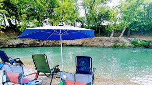 A river with chairs and an umbrella on a pebbled shore. - Charming Cabin on the San Marcos River with large back screened-in deck! - L2 (Kingsbury)