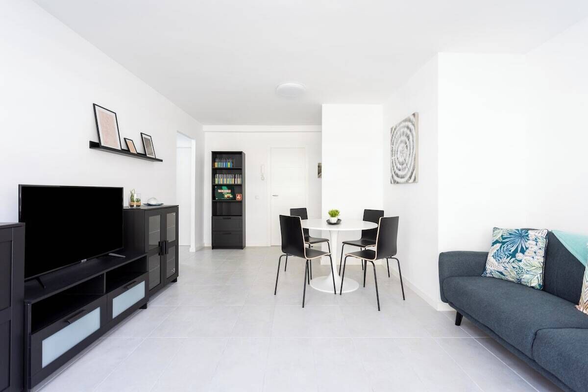 A modern living room with a flat-screen TV, a black entertainment unit, a dining table with chairs, and a grey sofa with a decorative pillow.