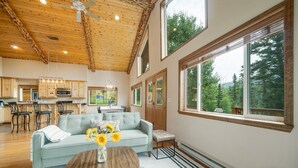 A spacious living area with a light blue sofa, a wooden coffee table, and a vase of sunflowers. There are large windows offering a view of trees and a kitchen area with wooden cabinets and a bar stool. - Welcome to Lemon Lodge! Relax and unwind in this private and secluded cabin.  (Durango)